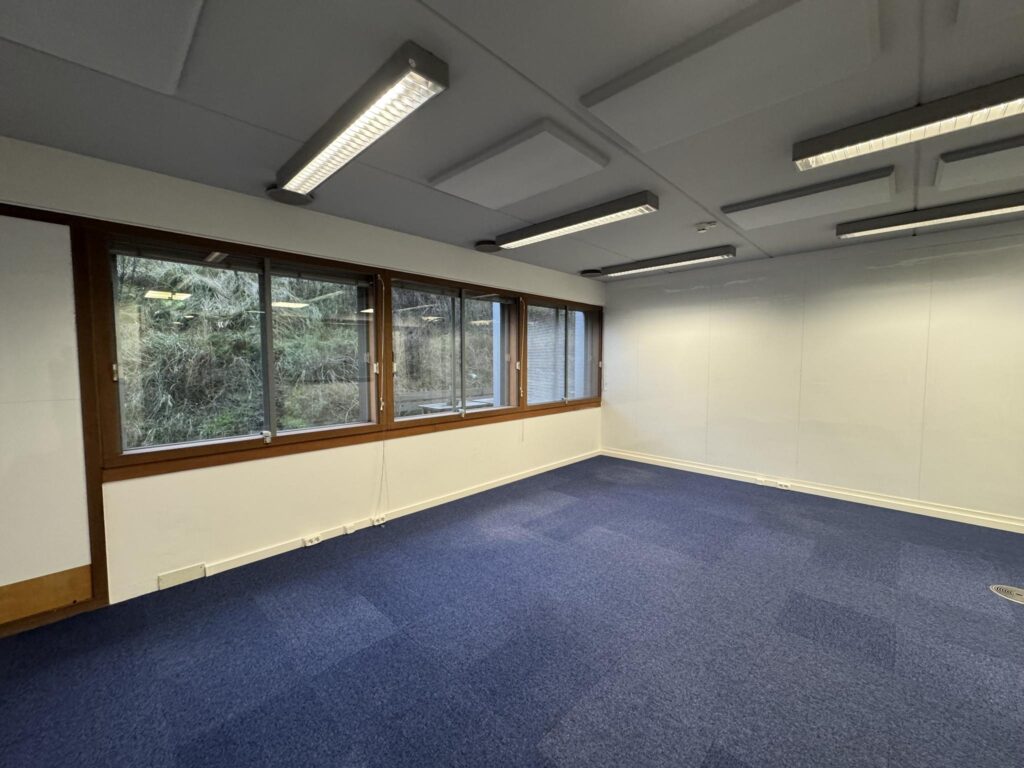 Empty office space with blue carpet, white walls, and large windows at De Ruijterkade.